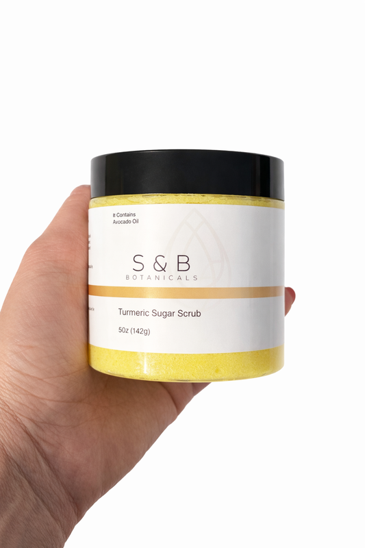 Hand holding S&B Botanicals Turmeric Sugar Scrub jar against white background showing full product label, 5oz natural brightening scrub with avocado oil and organic ginger root