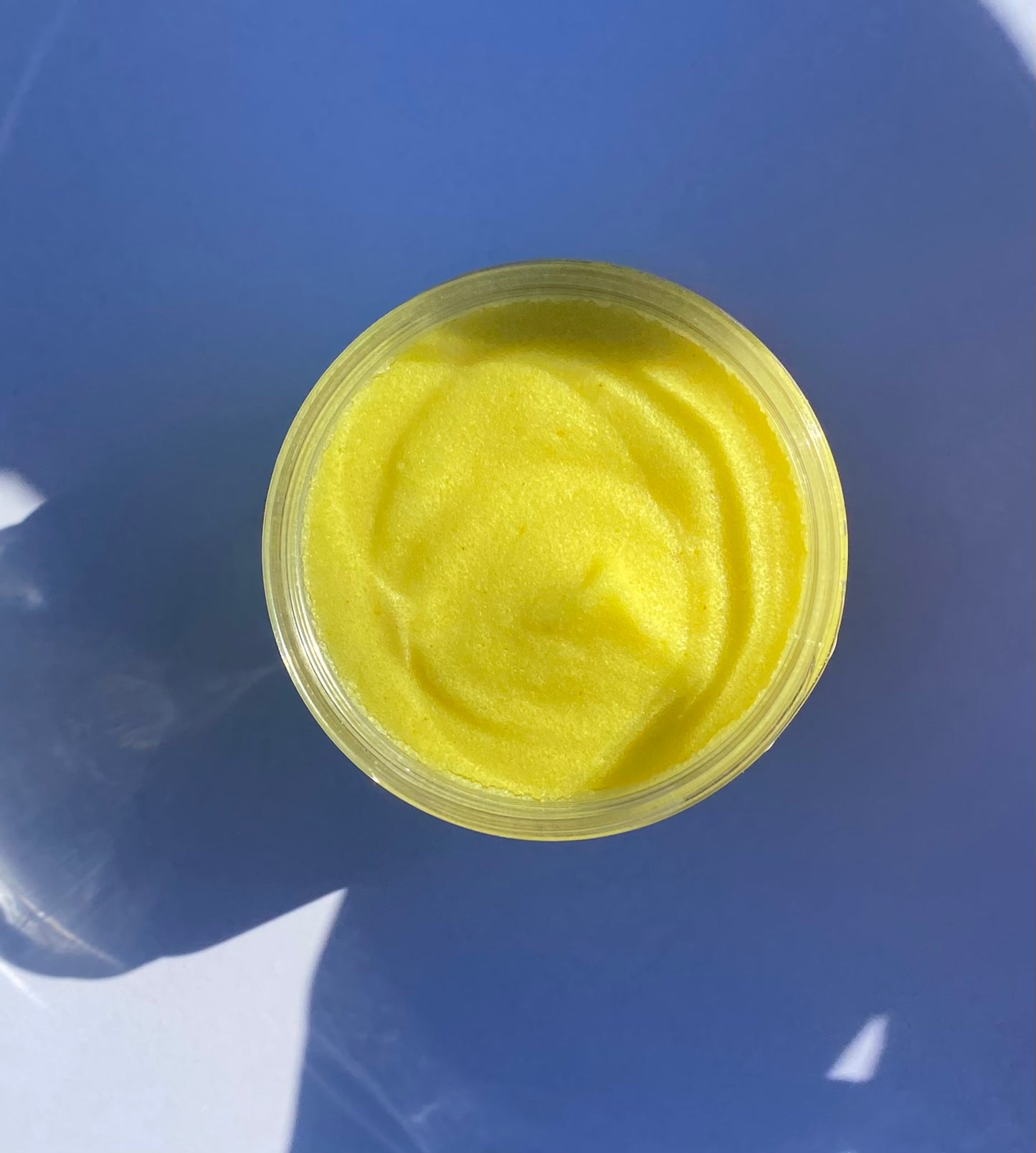 Overhead flat lay of open S&B Botanicals Turmeric Sugar Scrub jar on blue background showing smooth golden yellow sugar scrub texture, small batch natural exfoliating body scrub