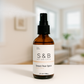 S&B Botanicals Room Spray amber glass bottle displayed on white surface in bright minimalist living room, 2oz water-based phthalate-free fragrance spray for any space