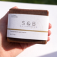 Hand holding S&B Botanicals Toasted Matcha Latte Soap Bar in natural sunlight outdoors, showing full labeled packaging with coconut milk and ceremonial matcha