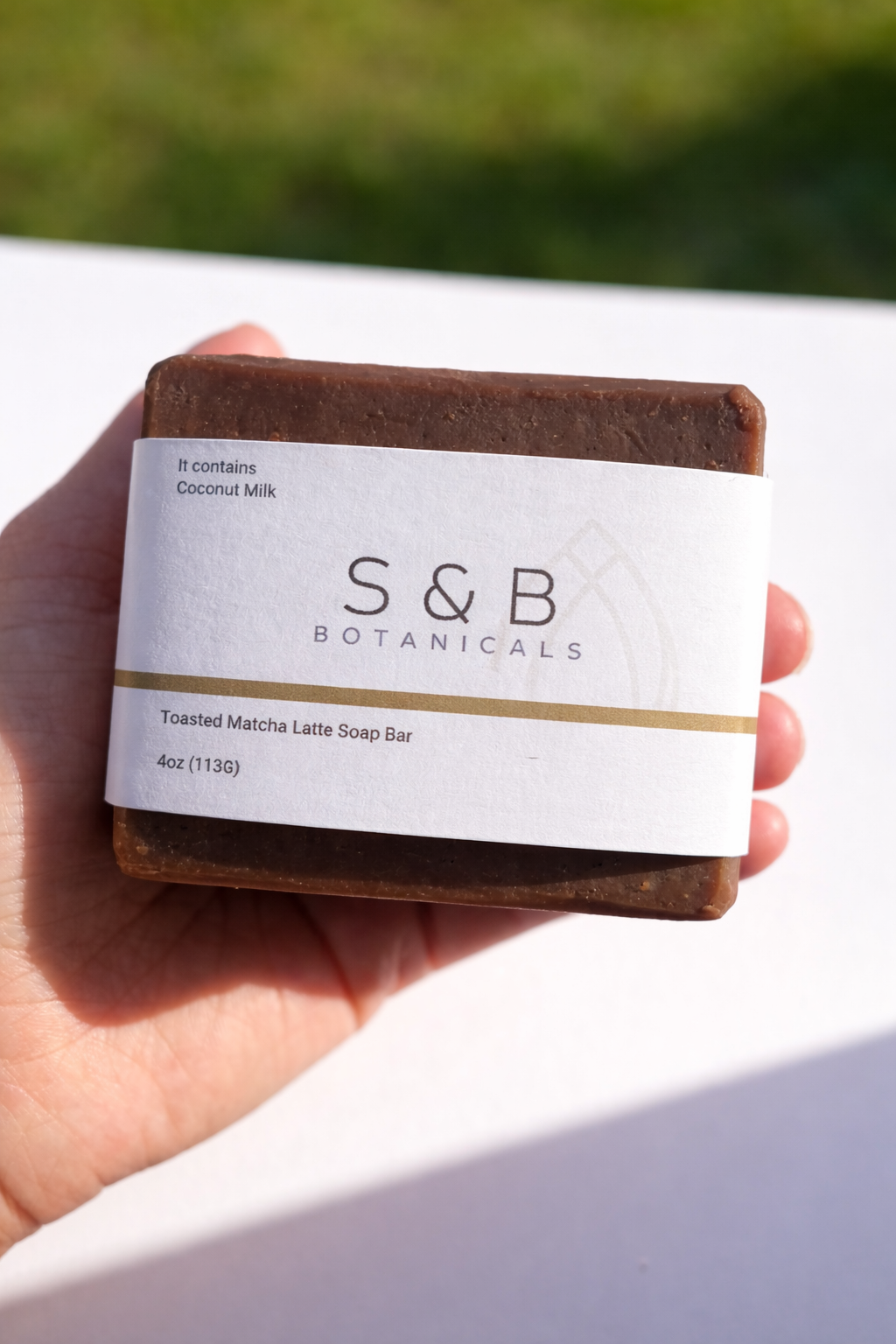 Hand holding S&B Botanicals Toasted Matcha Latte Soap Bar in natural sunlight outdoors, showing full labeled packaging with coconut milk and ceremonial matcha
