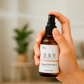Hand holding and spritzing S&B Botanicals Room Spray amber glass bottle in bright home setting with greenery in background, clean phthalate-free fragrance mist for bedrooms, linens, and more​​​​​​​​​​​​​​​​